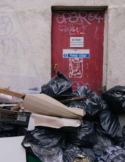Two large black plastic bags filled with waste material are situated on the pavement at the edge of a street, near a black metal fence and a hedge with dense, dark foliage. The bags appear to contain